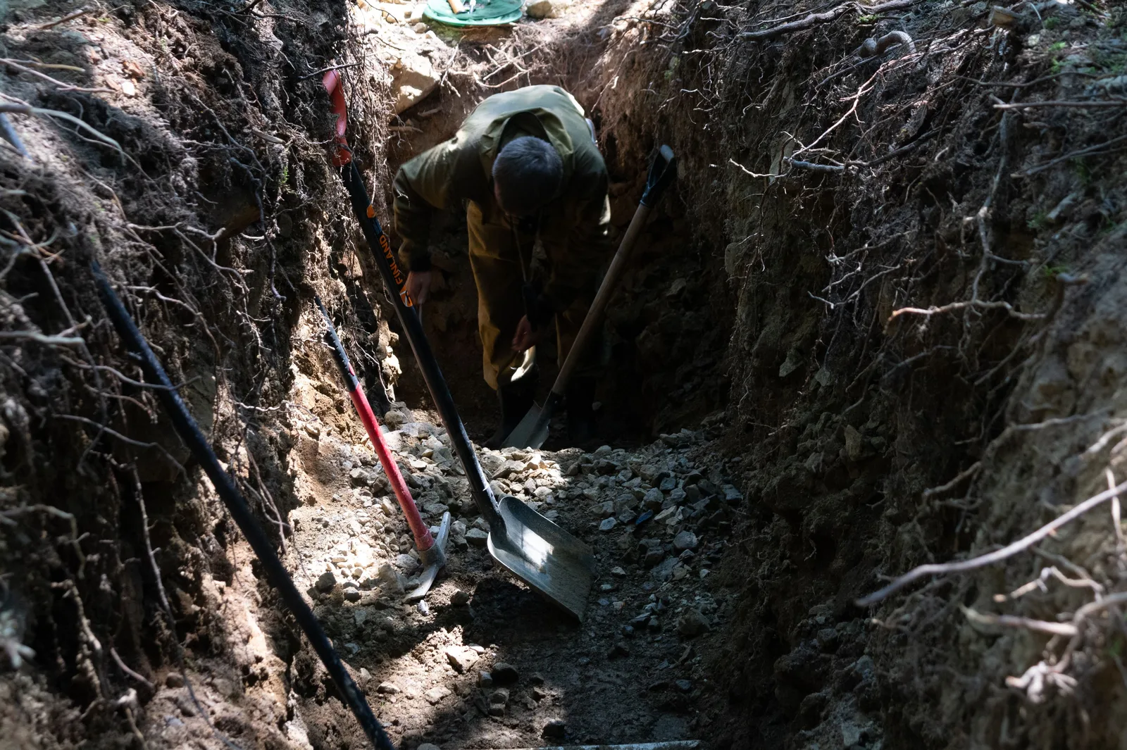 A person digging a trench outside, surrounded by tools and natural surroundings. Patio renovation regrets: the skipped test hole and the vague allowance